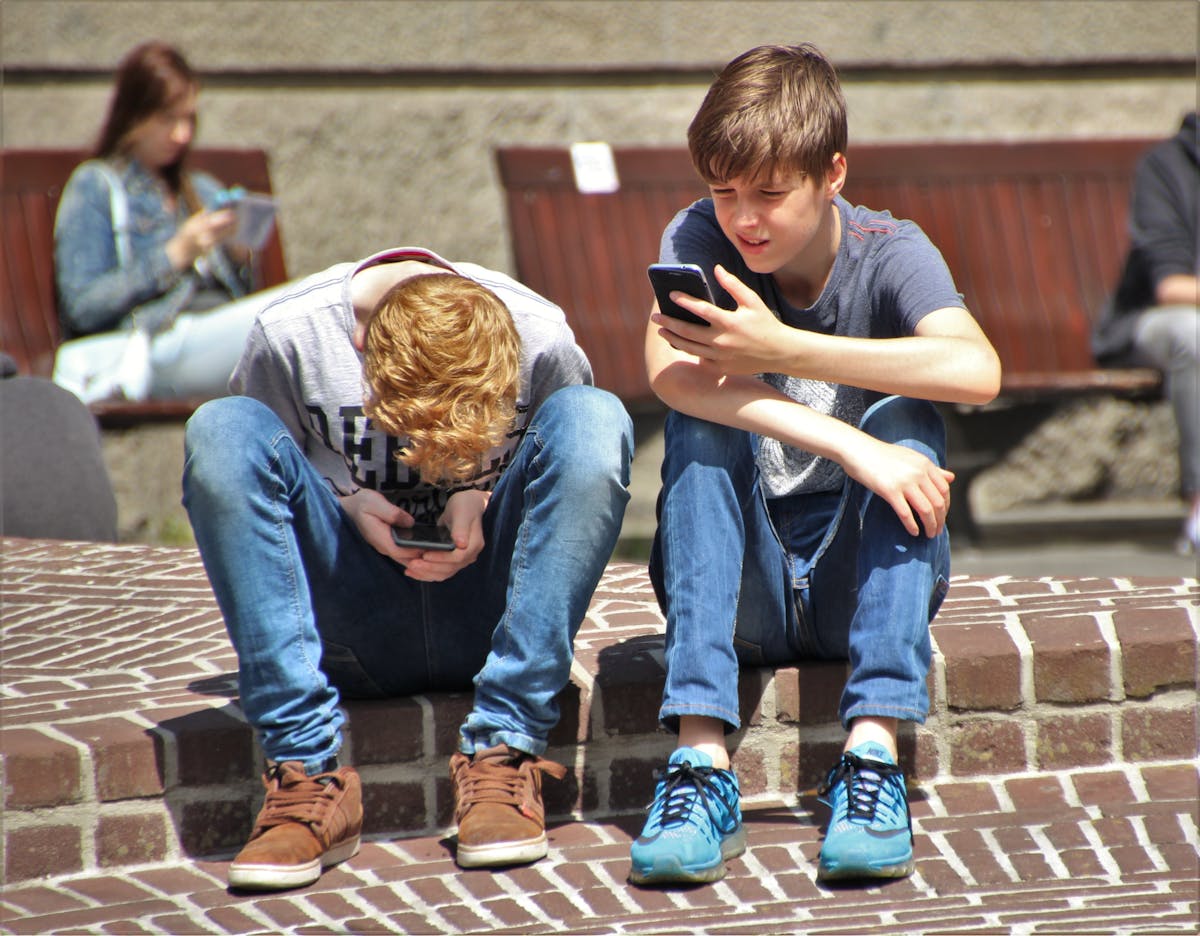 2 boy sitting on brown floor while using their smartphone near woman siiting on bench using smartphone during daytime 2 boy sitting on brown floor while using their smartphone near woman siiting on bench using smartphone during daytime