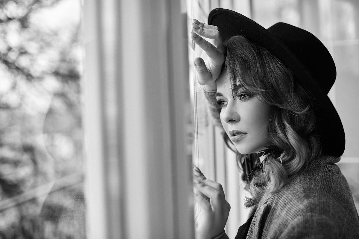 grayscale photo of woman in black hat leaning on glass window