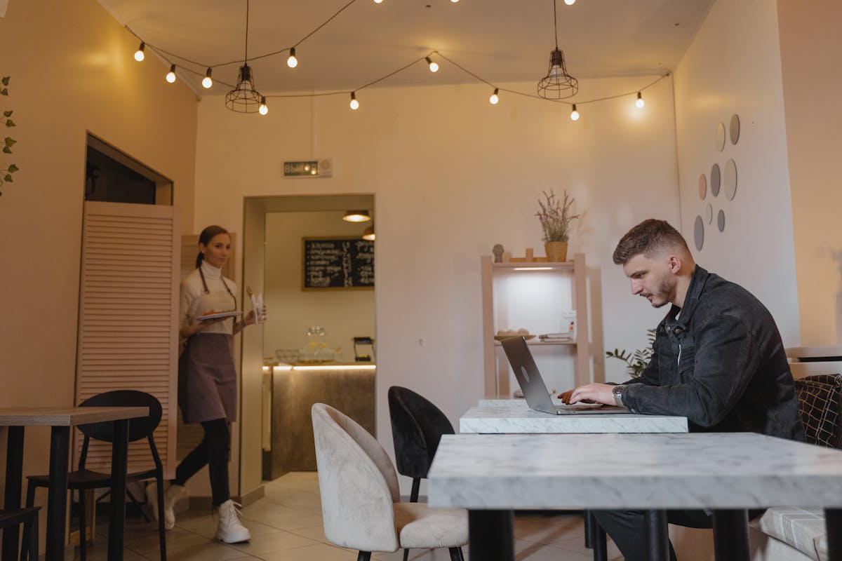man in black jacket using laptop inside a cafeteria with a woman serving food