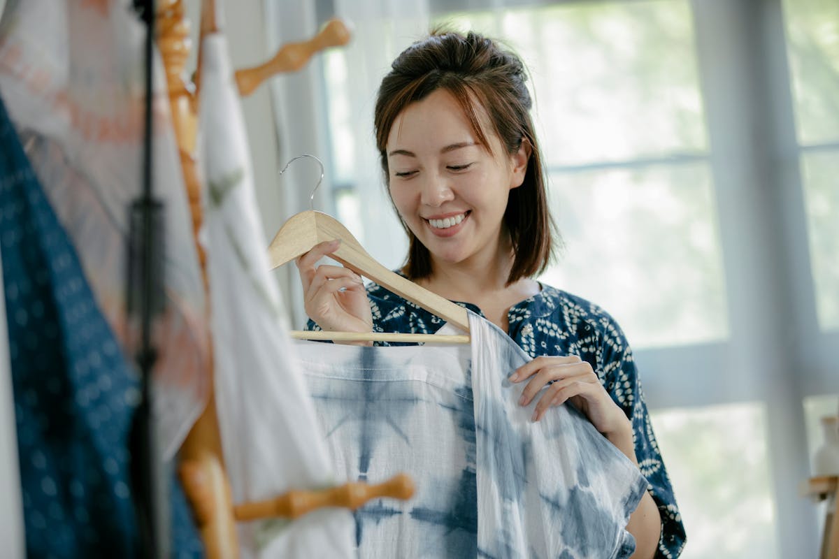 positive asian woman hanging clothes on hanger in workshop